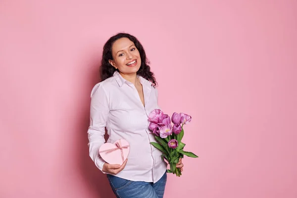 Stock image Attractive multi-ethnic positive pregnant woman, expressing happiness expecting for baby, posing with bunch of tulips and heart shaped gift box, smiling looking at camera, isolated on pink background.