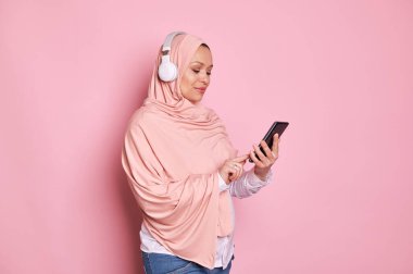 Pembe tesettürlü hamile bir kadın, akıllı telefondan mobil uygulamaları kontrol ediyor, çalma listesini kaydırıyor ve pembe arka planda izole edilmiş, kablosuz kulaklıklardan müzik dinliyor. Boşluğu kopyala