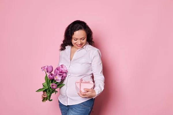 stock image Beautiful smiling pregnant woman posing with heart shaped pink gift box and bouquet of tulips over isolated pink background with copy advertising space. Happy Mothers Day. International Womens Day