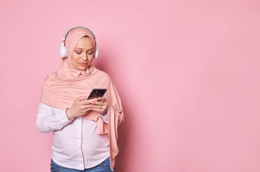 Ortadoğulu güzel, hamile bir kadın pembe tesettür ve kablosuz kulaklık takıyor, pembe arka planda akıllı telefon kullanıyor. İnsanlar ve teknoloji. Mobil uygulamalar. Çevrimiçi iletişim