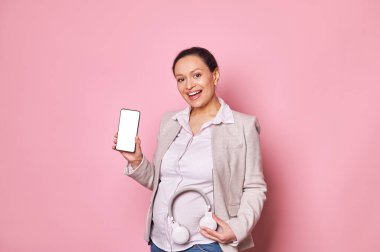 Karnına kulaklık dayamış hamile bir kadın, akıllı telefonunda beyaz boş bir ekran ve reklam alanı gösteriyor. Mobil uygulamalarınızı takmak için. Kameraya bakıp gülümsüyor.