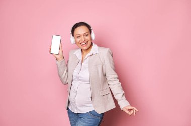 Mutlu gülümseyen hamile kadın kulaklıkla müzik dinler, pembe arka planda dans eder, elinde mobil uygulama için fotokopi alanı olan beyaz boş dijital ekranlı akıllı telefon tutar. - İnsanlar. Teknoloji