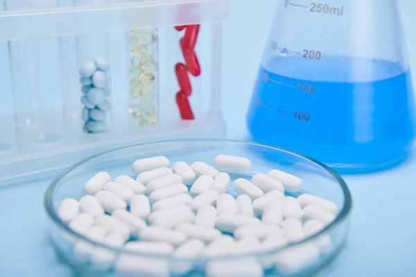 Selective focus on pile of white medical pills in petri dish against labware with capsules and blue solution, isolated on blue background. World Health Day. Pharmaceutical industry. Clinical research