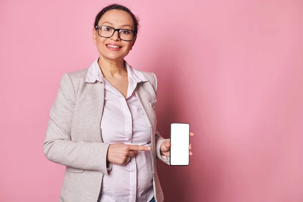 stock image Happy pregnant woman showing you a smartphone with blank white digital screen with mockup for mobile application, smiling looking at camera, isolated pink background. Copy space for advertising text