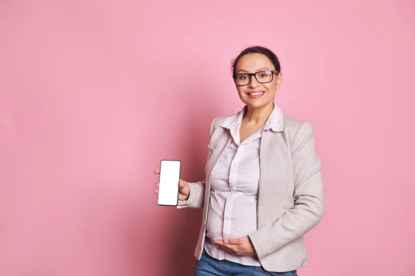 Middle Eastern pregnant pretty woman in eyeglasses, touches belly, smiles, shows cell phone with empty white digital screen, free ad space for insert your mobile application, isolated pink background