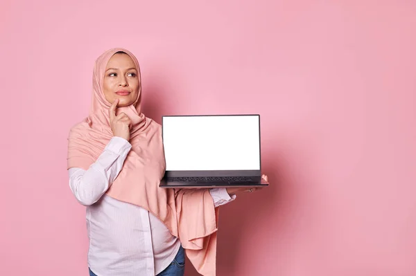 ピンクのヒジャーブ州に住むイスラム教徒の妊婦は 白い空白の画面を持ち 慎重に脇を見て ビジネスプロジェクトの推論 彼女の産休の計画 孤立したピンクの背景 — ストック写真