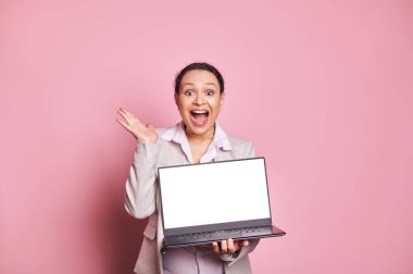 Şaşırmış etnik bir kadın, ağzı açık, mutlu olumlu duyguları ve şaşkınlığı ifade ediyor, dizüstü bilgisayarını beyaz boş ekranlı, mobil uygulamalar için fotokopi alanı olan, izole edilmiş pembe arka plan