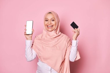 Pembe tesettürlü gülümseyen Ortadoğulu Müslüman kadın, kameraya gülümsüyor, elinde siyah kredi kartı ve beyaz beyaz ekranlı akıllı telefon, izole pembe arka plan. Mobil uygulama için reklam alanı, metinler