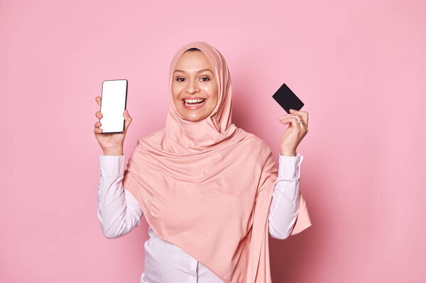 Smiling Middle Eastern Muslim woman in pink hijab, smiling looking at camera, holding black credit card and smartphone with white blank screen, isolated pink background. Ad space for mobile app, texts