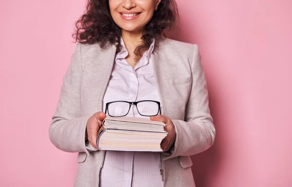 Details on stylish eyeglasses on a book in hands of blurred pretty woman, school teacher, or librarian smiling a cheerful toothy smile, isolated on pink background. World Book Day and Teachers Day