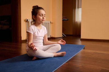 Kendine güveni tam, 5-6 yaşlarında beyaz tişörtlü, gri bacaklı güzel bir kız çocuğu. Lotus pozunda spor minderinde oturuyor. Yoga yapıyor, rahat bir evde meditasyon yapıyor..