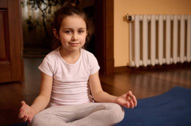 Güzel Avrupalı rahat küçük kız spor üniformalı, kameraya bakıp yoga minderinde meditasyon yaparken gülümsüyor..