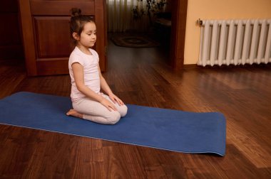 Sevimli beyaz çocuk meditasyon yapıyor. Yoga yaparken gözleri kapalı bir şekilde spor minderinin üzerinde oturuyor.