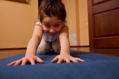 Ön cepheden yakın çekim. Sevimli 5-6 yaş arası beyaz bir kız çocuğu çocuk pozuyla vücudunu esnetiyor, içerde yoga minderinde egzersiz yaparken kameraya bakıp gülümsüyor. Spor. Fitness. Sağlıklı alışkanlıklar