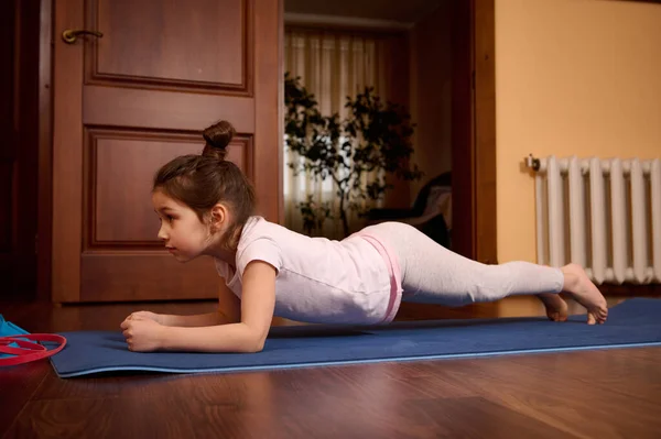 Determined Caucasian little girl doing push ups, press ups, four limbed staff exercise, staying in plank on a blue fitness mat while practicing stretching and yoga in cozy wooden homely atmosphere