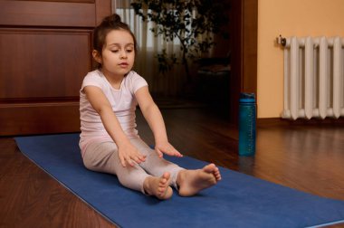 Azimli küçük kız 5-6 yaşlarında, yoga minderinde çıplak ayak egzersiz yapıyor, ısınıyor, vücudunu esnetiyor, rahat ahşap atmosferde jimnastik yapıyor. Spor ve insanlar konsepti