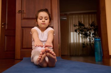 Sevimli çocuk egzersiz yapıyor, mavi bir spor paspasın üzerinde çıplak ayakla oturuyor evde yoga yapıyor ve egzersiz yapıyor. Spor. Fitness. Jimnastik. Aktif ve sağlıklı yaşam tarzı kavramı