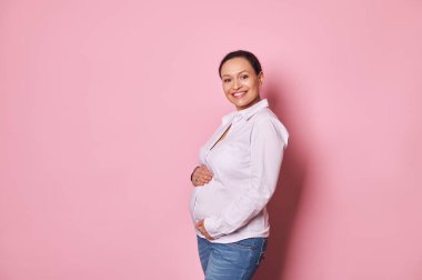 İzole edilmiş portre pembe arka plan. Çok ırklı hamile bir kadın, karnına dokunuyor, ilk bebek tekmelerinin tadını çıkarıyor, kameraya bakıp gülümsüyor. Bebek bekleyen oldukça ağır bir kadın. Sağlıklı hamilelik kavramı