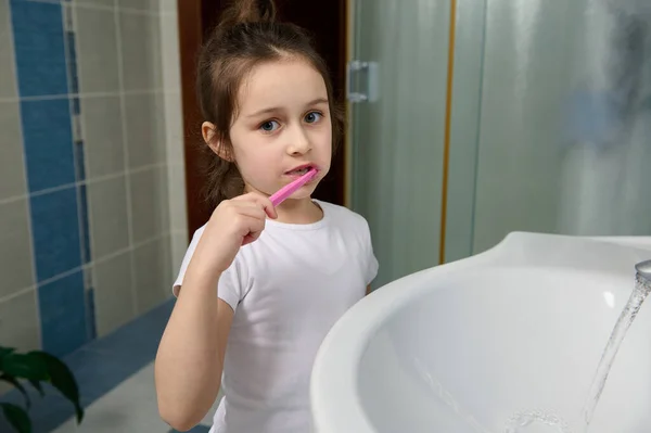 Morning daily routine concept. Dental care and oral hygiene for healthy white baby teeth. Adorable kid girl standing by white ceramic sink in light blue bathroom, brushing teeth and looking at camera.