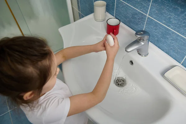 Hands hygiene and virus infections prevention. Overhead view of a conscious little child girl, washing her hands with soap bar at home bathroom. The concept of hygiene and clean hands. Selective focus