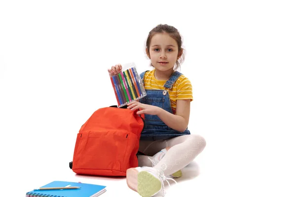 可爱的白人幼儿 一年级的小女孩坐在白色的背景上 把彩色的毛笔放进她的橙色背包里 微笑着看着相机 全长肖像 — 图库照片