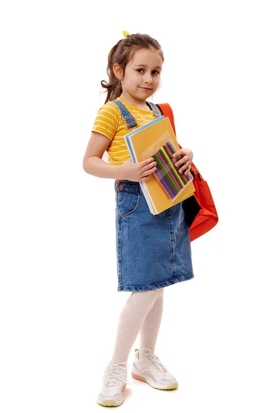 6歳の子供の完全な長さの肖像画 黄色のTシャツと青のデニムのサンドレスのスマート女子高生は バックパックや学校用品を運ぶ 笑顔かわいいカメラを見て 隔離された白い背景 — ストック写真