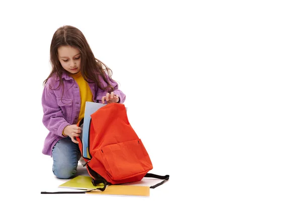 カジュアルなデニムと紫のシャツを着て 彼女のオレンジ色のバックパックに教科書や学校用品を折り 白い背景に隔離されたコーカシアの愛らしいスマート女子高生のフルサイズの肖像画 — ストック写真
