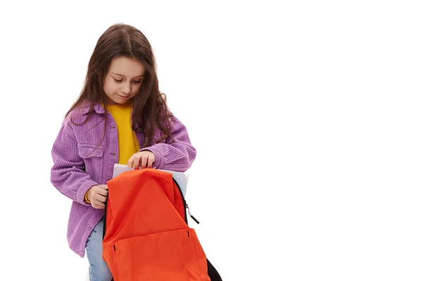 可爱的女学生把学习用品包在背包里 坐在孤立的白色背景上 脸上挂着可爱的笑容 穿着紫色天鹅绒衬衫 复制广告空间 回到学校的概念 — 图库照片
