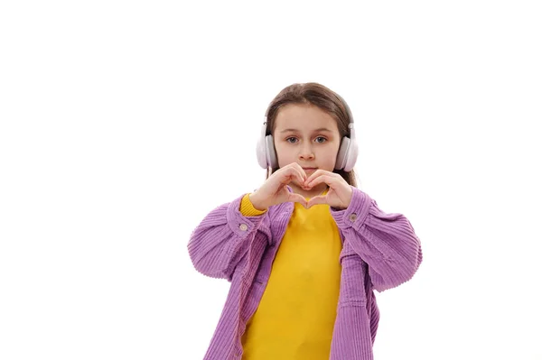 ヘッドフォンで幸せな白人の子供の女の子 彼女の指から作られた心の形を示します 愛と同情を表現 カメラを見て 白い背景に隔離されました 国際子どもの日のコンセプト — ストック写真