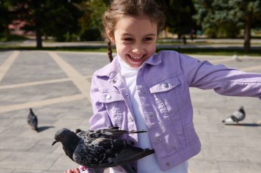Neşeli küçük beyaz kız çocuğu, güneşli bir bahar günü parkta uçan kuş sürüsünü beslerken kolunda oturup gülümseyen ve mutluluğunu ifade eden bir güvercine seviniyor.