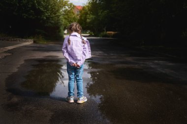 Kotlu ve mor ceketli küçük bir kız, yağmurdan sonra elleri çapraz duran küçük bir su birikintisi ve kırsal yolda yürürken sudaki yansımasına bakıyor. Arka görünüm