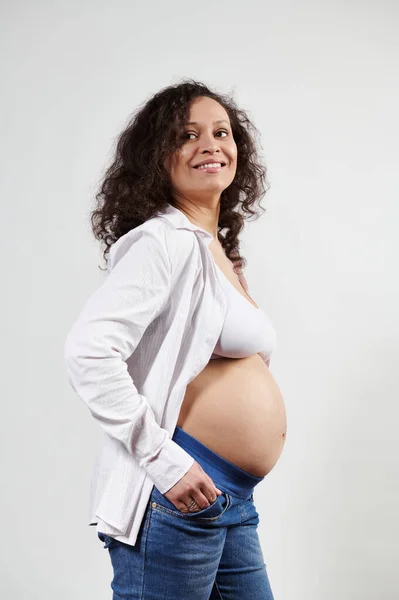 セクシーな巻き毛の妊婦で妊娠6ヶ月目 青いジーンズと白い下着で 脇を見て笑って ポケットに手を入れて 孤立した白い背景に裸の腹でポーズ — ストック写真