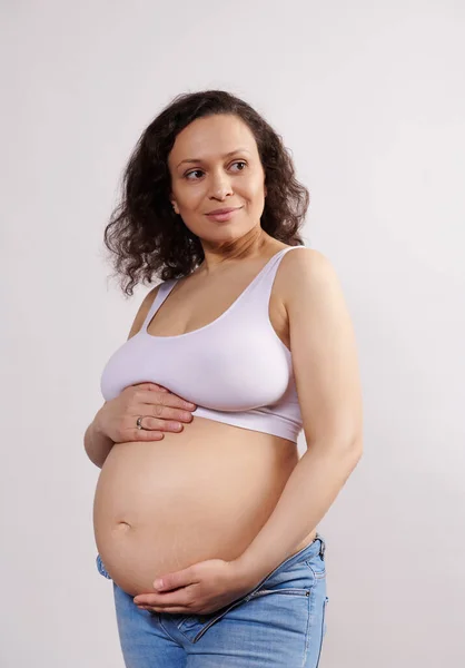 拉丁美洲35岁至39岁的年轻女子 自信而快乐 面带微笑地将目光移开 爱抚怀孕的腹部 快乐而无忧无虑地怀孕 与白色背景隔离 — 图库照片