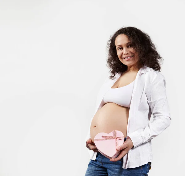 面带微笑的卷发黑发画像 孕妇在腹部附近抱着粉色心形礼品盒 与白色背景隔离 母亲节快乐 无忧无虑的怀孕和分娩概念 — 图库照片