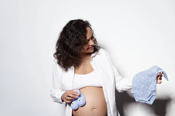 白い背景に隔離された 妊娠時間で物事を準備し 将来の新生児のための青いボディスーツと編んだ赤ちゃんのおっぱいを保持し 大きな腹を持つ現在髪の若い大人の妊婦 — ストック写真