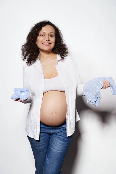 将来の新生児のために可愛い青い赤ちゃんのおっぱいとボディスーツを持っている陽気な幸せな巻き毛の妊婦は 白い背景に隔離されたカメラで笑っています 出産だ 妊娠第2期 — ストック写真