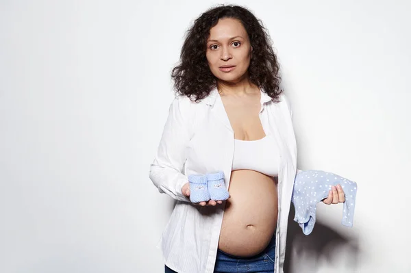 中年齢の多民族妊娠可愛い女性の第二妊娠の三半期の肖像画のショット カメラを見て 白い背景に青い新生児の体のスーツと赤ちゃんの靴でポーズ 広告スペースのコピー — ストック写真