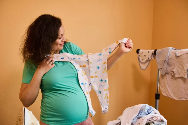 Stock image Multi ethnic middle aged beautiful curly haired woman, pregnant mother in home wears, holding and looking at small cute baby outfit, preparing equipment for newborn childbirth. Pregnancy 3d trimester