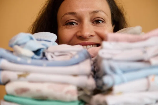 Close-up cheerful happy woman smiling cutely looking at camera through a stack of ironed laundered clean newborn clothes. The concept of maternity, pregnancy and preparation for the birth of a child