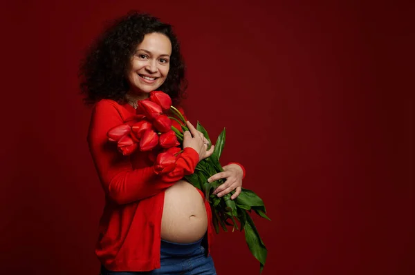 美しいアフリカ系アメリカ人の妊婦がカメラを見て大笑いし 孤立した色の背景に赤いチューリップの束で裸の腹をポーズします 幸せな母と女性の日の概念 広告スペース — ストック写真