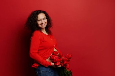 Mutlu kadınlar ve anneler günü konsepti. Güzel, etnik, esmer, hamile bir kadın. Elinde bir sürü kırmızı lale var. Kameraya bakıp olumlu duygular ifade ediyor.