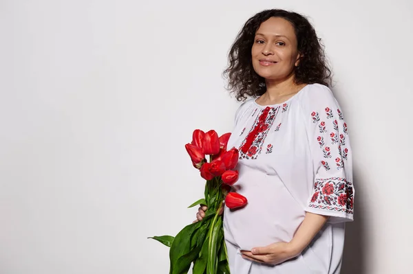 幸せな楽しい妊娠中の女性は 赤いチューリップの花束を持って 彼女の腹を愛撫 隔離された白い背景にカメラを見て 伝統的なウクライナの刺繍白いドレスを着て — ストック写真