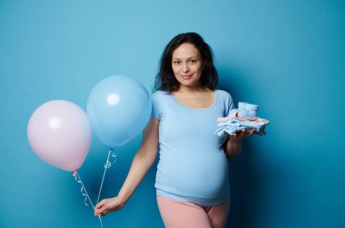 30 'lu yaşlarda güzel hamile bir esmer kadın, kameraya bakıp gülümsüyor, elinde ütülenmiş yeni doğan giysileri, bebek paçaları ve mavi arka planda izole edilmiş pembe mavi hava balonları tutuyor. Hamilelik. Cinsiyet partisi