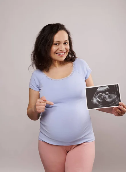 stock image Happy adult pregnant woman, smiling looking at camera, showing her newborn baby sonography and pointing at her belly over isolated isolated white background. Pregnancy. Ultrasound scan image of a baby