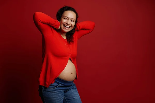 卷发的怀孕的成年妇女笑着 在孤立的红色背景上摆出赤裸的腹部 享受快乐无忧无虑怀孕的美妙时刻 复制空间 大家好生活方式 — 图库照片