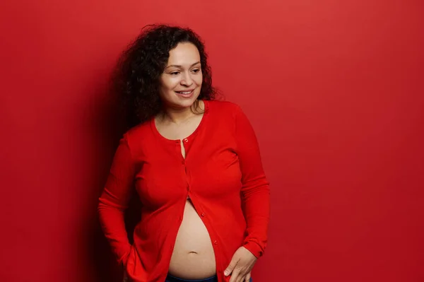 赤いボタンのないシャツの幸せな普及妊婦 優しく彼女の腹に触れ 思慮深く脇を見て 出産や妊娠についての推論は 孤立した赤の背景 スタジオポートレート — ストック写真