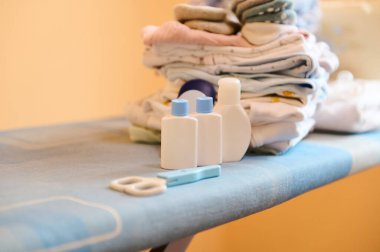 Yeni doğan bebek kozmetik ürünleri, tırnak makası ve ütü masasındaki temiz ütülenmiş giysilerin yakın plan çekimi. Modelleme. Çamaşır yıkamak, doğum hastanesi ve doğum için çanta hazırlamak. Uzayı kopyala
