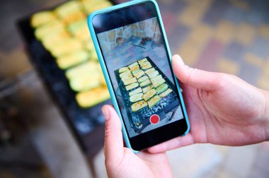 Cep telefonunu canlı izleme modunda tutan ve arka bahçedeki ızgarada mangal kömürü üzerinde ızgara yapan lezzetli salamura edilmiş sebzelerin fotoğraflarını çeken kadın eller. Yaz pikniği kavramı. Sakin ol.