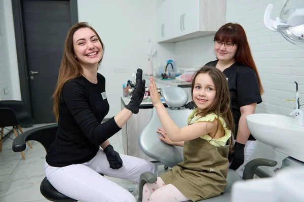 Little child girl smiles looking at camera, gives high five to her dentist hygienist doctor after teeth check up in a modern dental clinic. Pediatric aesthetic dentistry. Oral care and hygiene concept