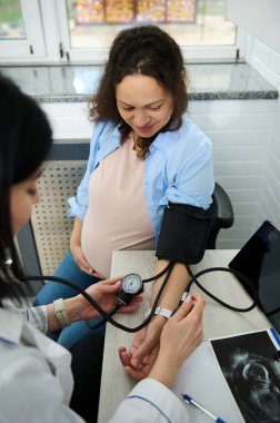 Mutlu hamile kadın hamilelik danışmanlığı için doğum hastanesinde ya da klinikte jinekoloğu ziyaret ediyor. Kadın doğum uzmanı, tonometreyle hamile bir kadının kan basıncını ölçüyor.